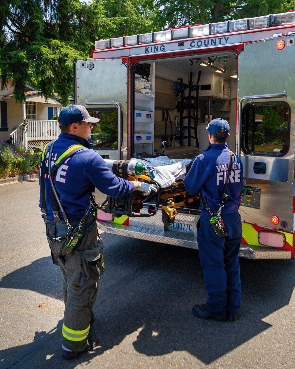 Two firefighters and an EMT load a stretcher into the back of a “King County” ambulance on a residential street in daylight.