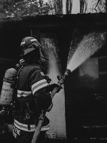 A black and white photo of a firefighter from the Fire Department spraying water on a house.