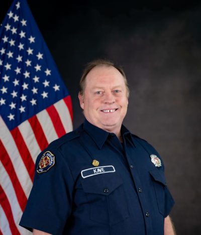 A firefighter in uniform standing in front of an american flag.