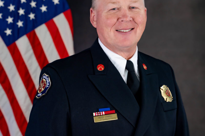 A man in a Valley Regional Fire Authority firefighter uniform standing in front of an american flag, representing the dedication and service of the Fire Department.