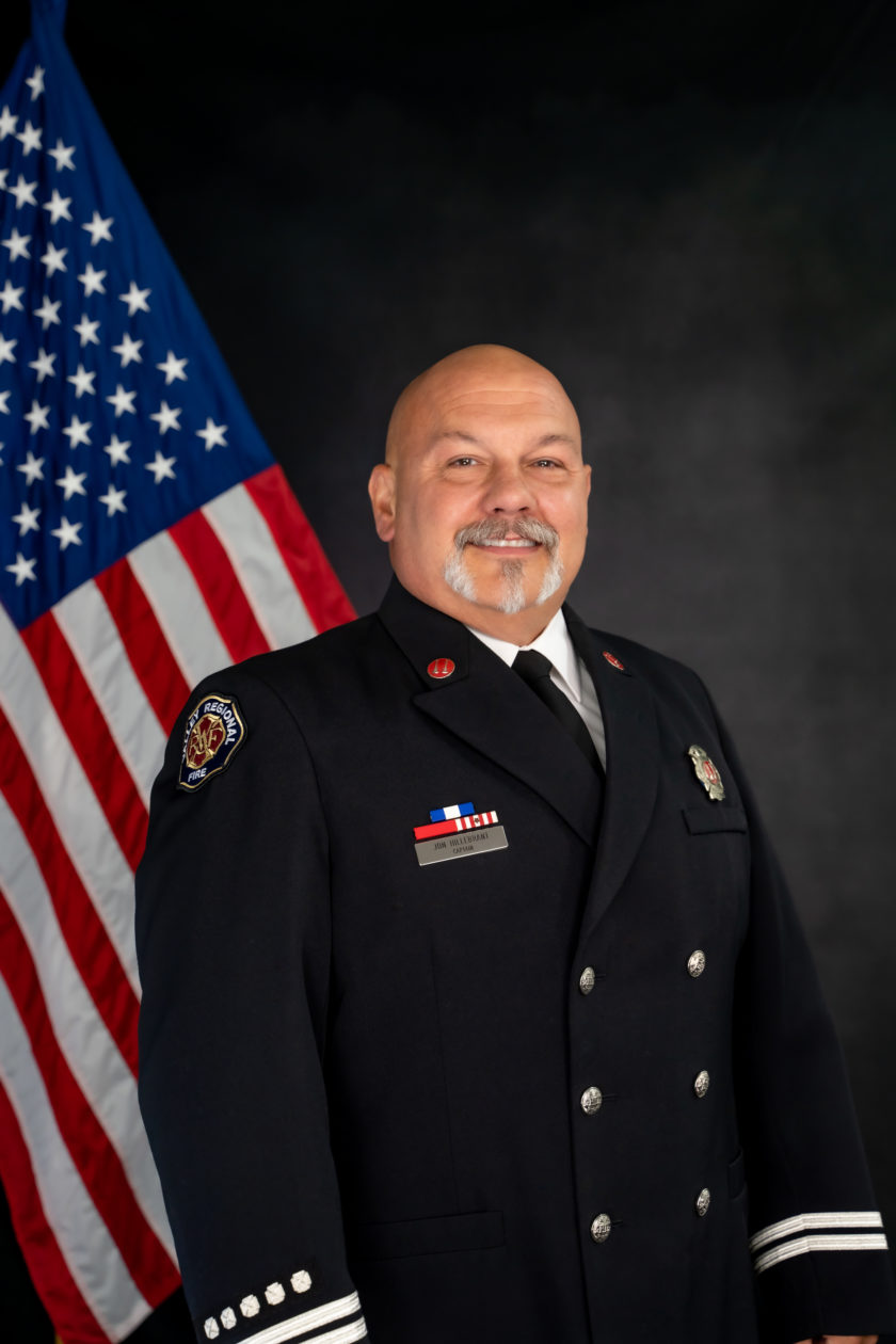 A man in a firefighter uniform standing in front of an american flag, representing the dedication and service of the Fire Department.