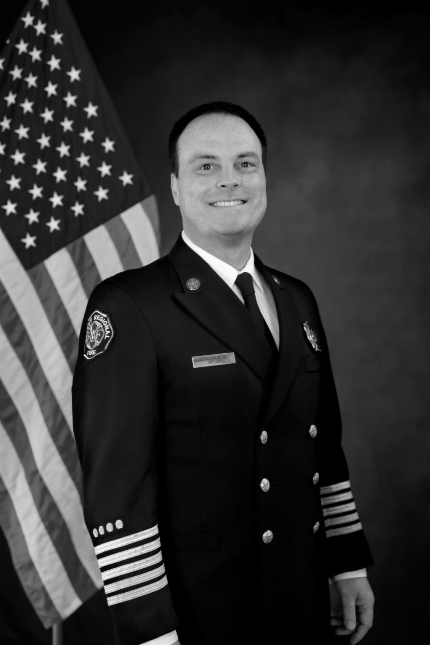 A black and white photo of a firefighter standing in front of an American flag, representing the bravery and dedication of the fire department.