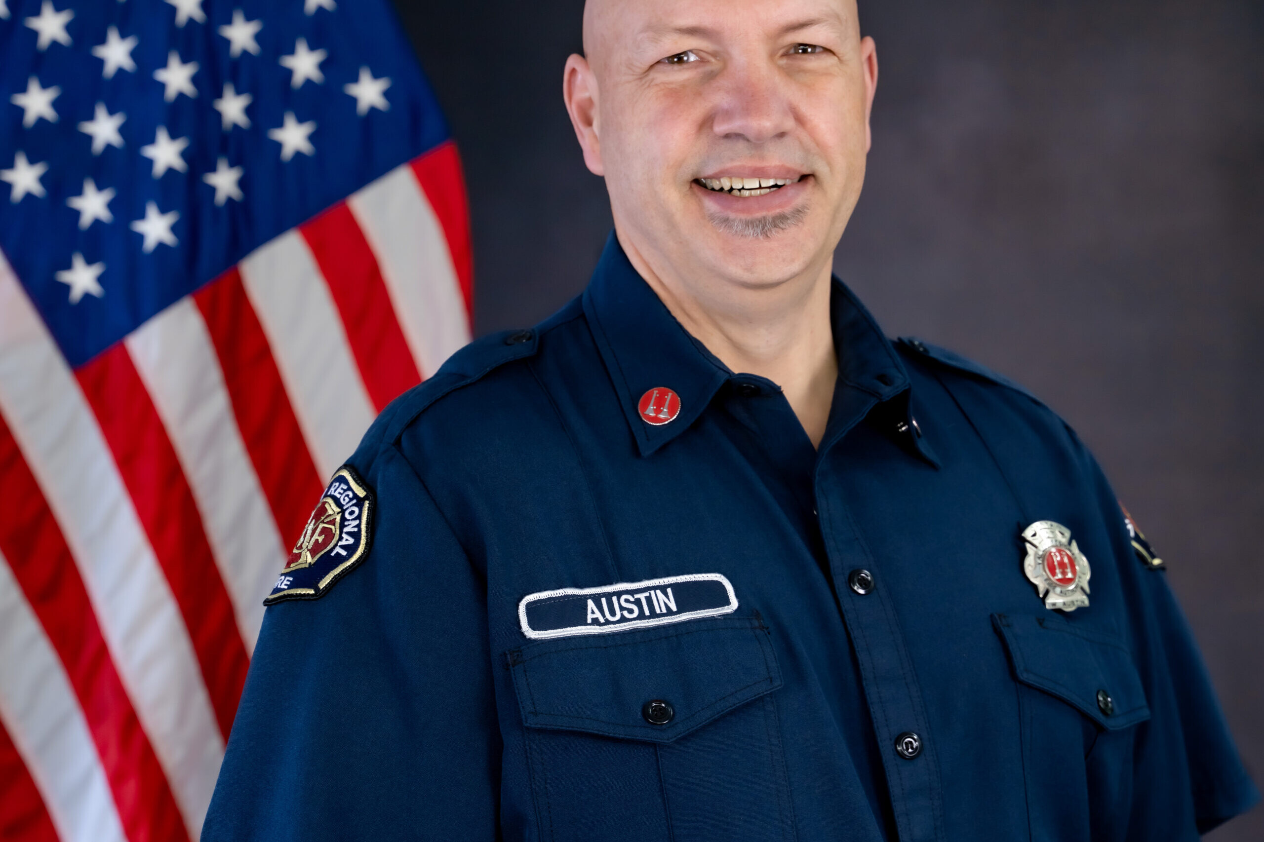 Captain Bill Austin, a bald man in uniform, retires after a distinguished career of service, pictured standing proudly in front of an American flag.