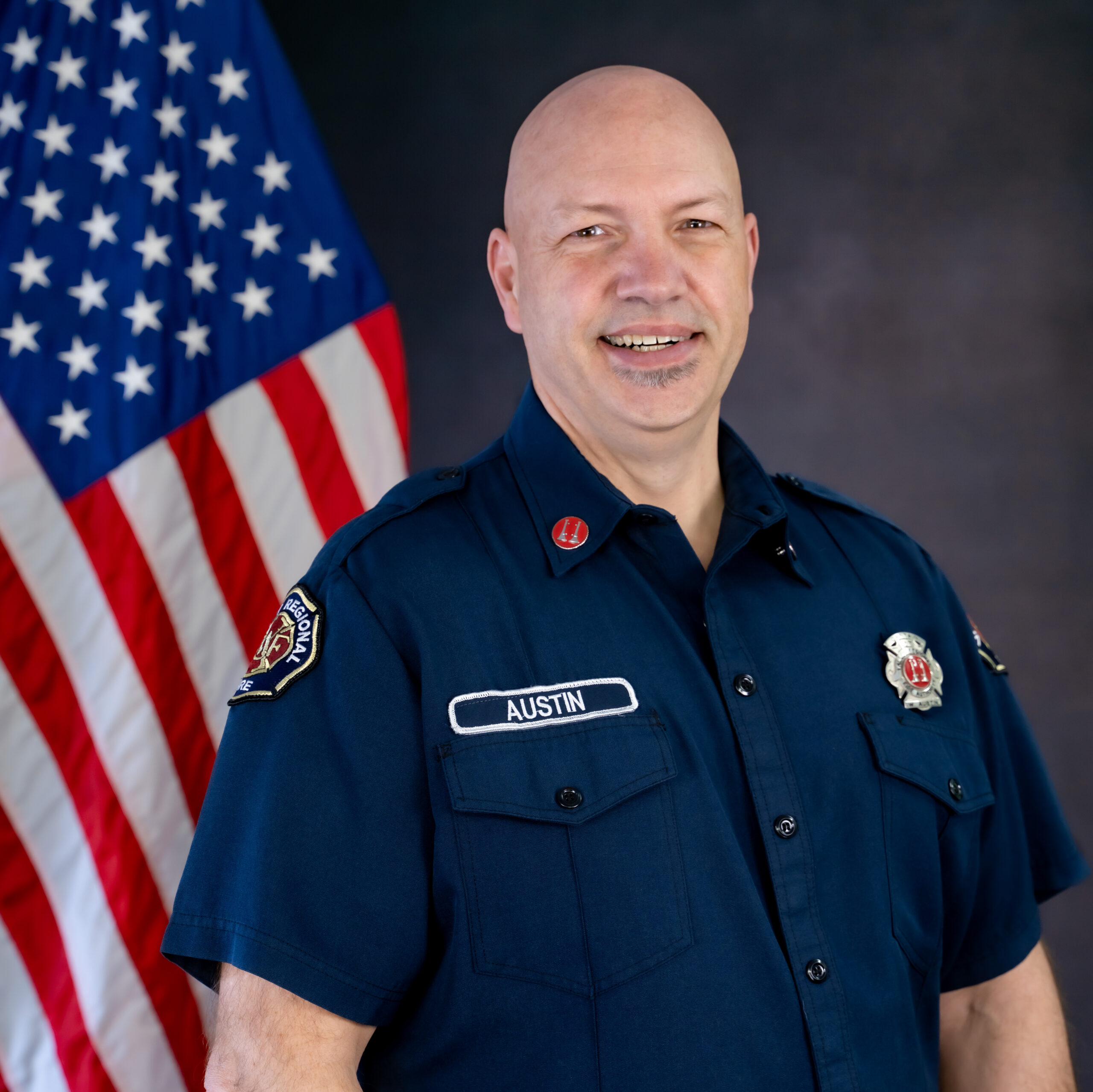 Captain Bill Austin, a bald man in uniform, retires after a distinguished career of service, pictured standing proudly in front of an American flag.