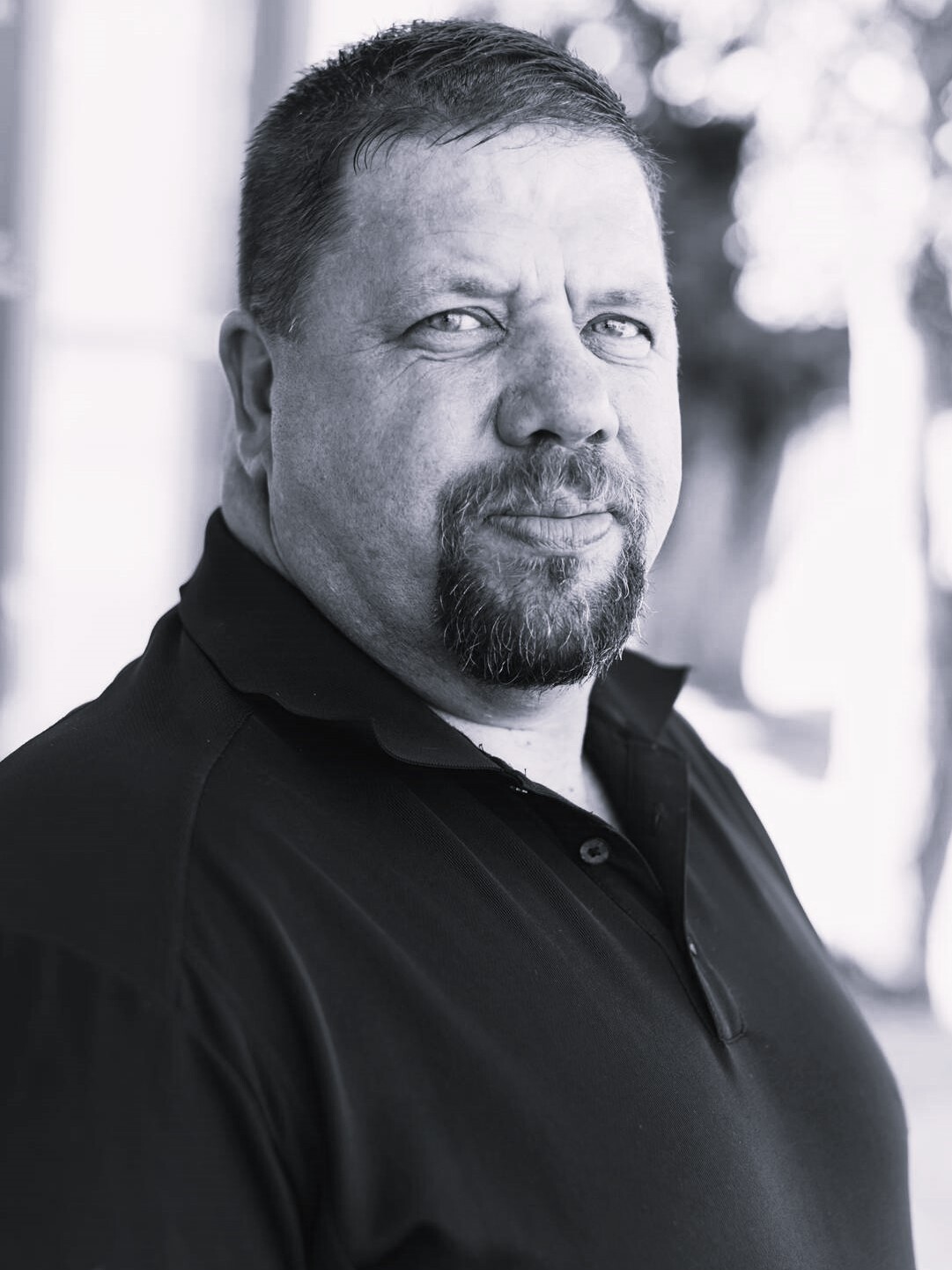 Black and white portrait of a middle-aged man with a beard, looking over his shoulder, wearing a black polo shirt.