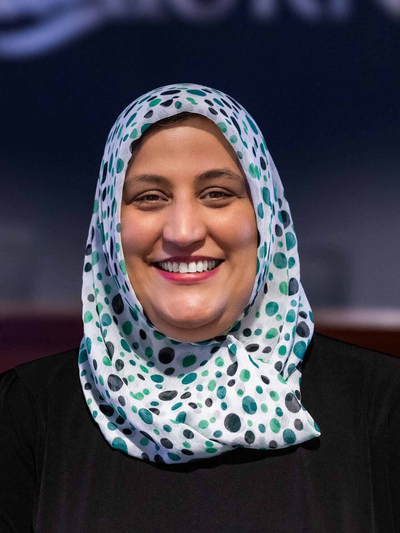 A woman wearing a white hijab with green and black dots and a black top smiles at the camera in an indoor setting at the Valley Regional Fire Authority.