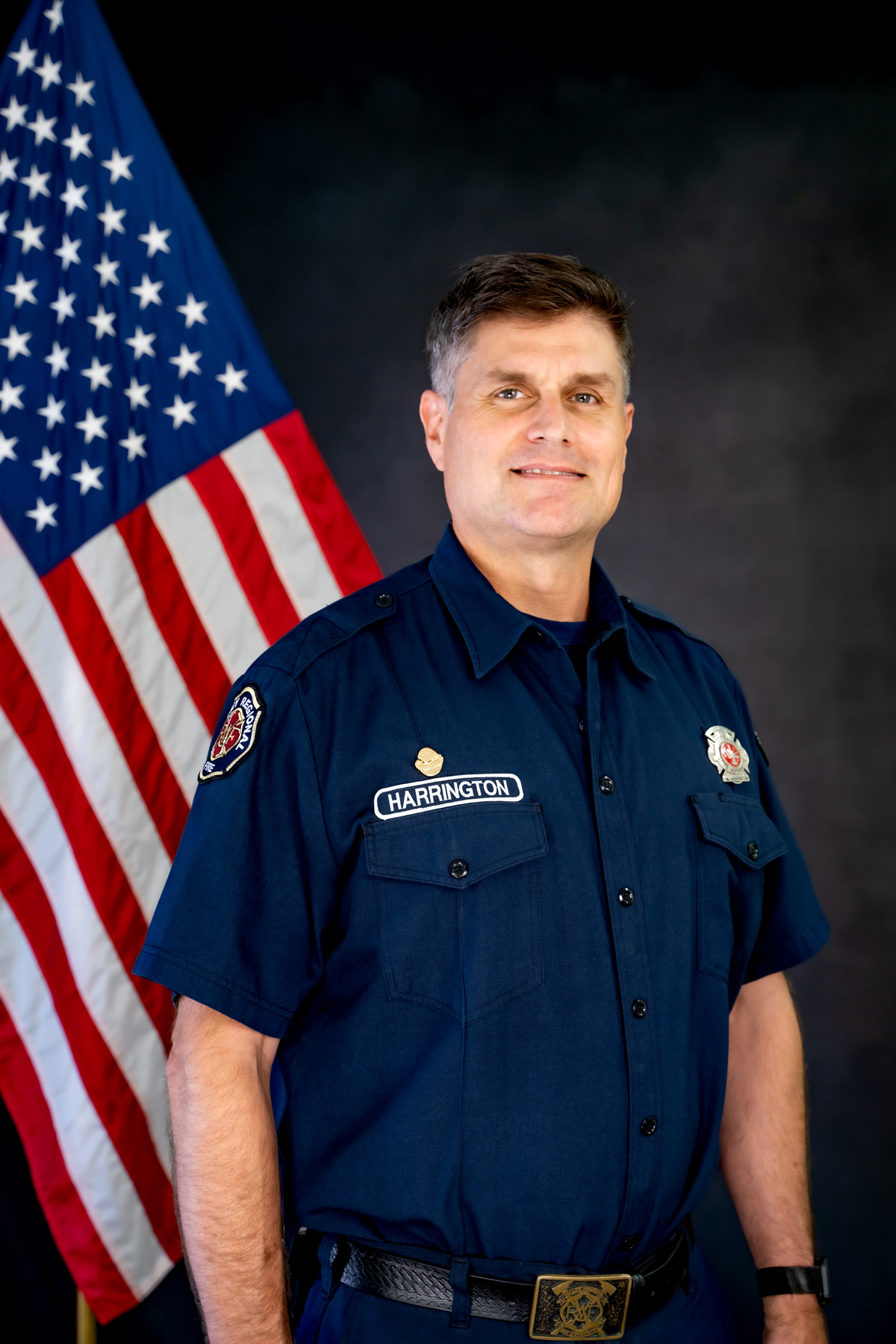 Deputy Fire Marshal Matt Harrington of the Valley Regional Fire Authority stands in a navy blue uniform before an American flag, looking at the camera with a neutral expression.