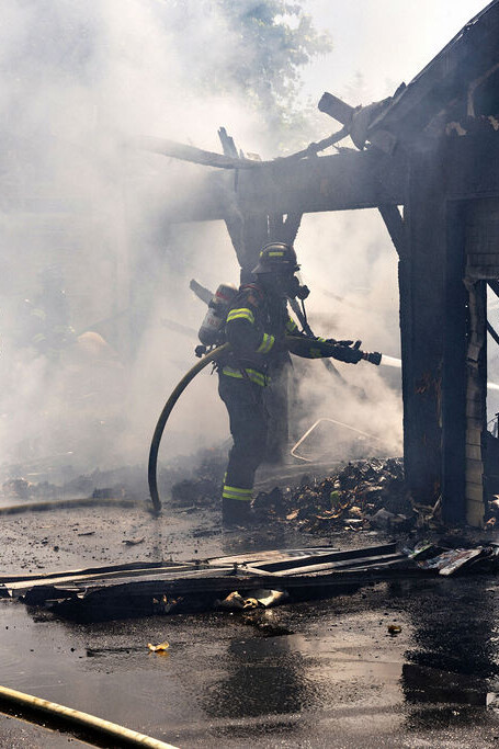 Three firefighters in protective gear work to extinguish a smoky fire in a partially destroyed building with debris and hoses on the ground.