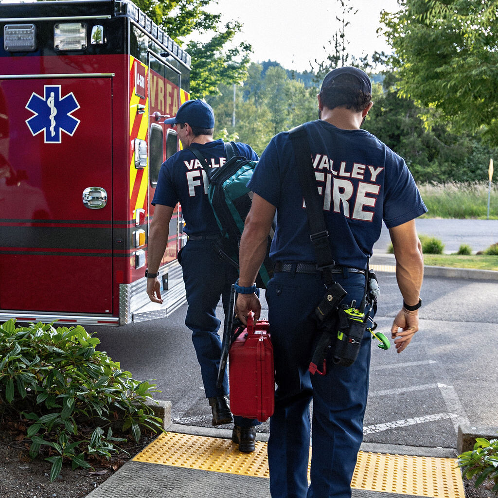 Two firefighters in uniform walk toward an ambulance, one carrying a red medical kit and the other carrying equipment, in a parking lot surrounded by greenery.
