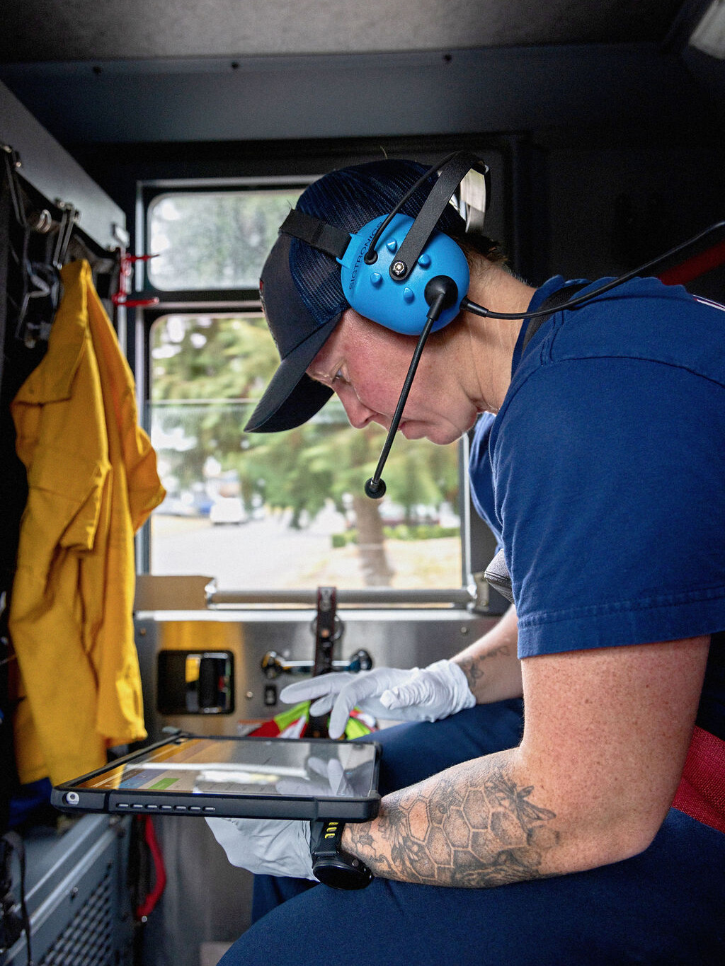 A person wearing a headset and gloves sits in a vehicle, looking at a tablet device, with emergency equipment visible nearby.