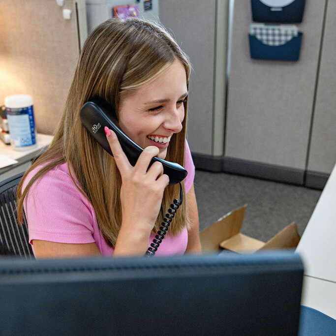 A woman sitting at an office desk smiles while talking on a corded phone in a cubicle workspace.