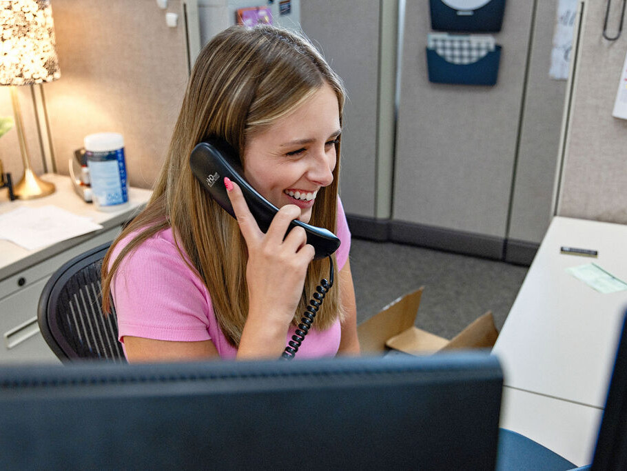 A woman sitting at an office desk smiles while talking on a corded phone in a cubicle workspace.