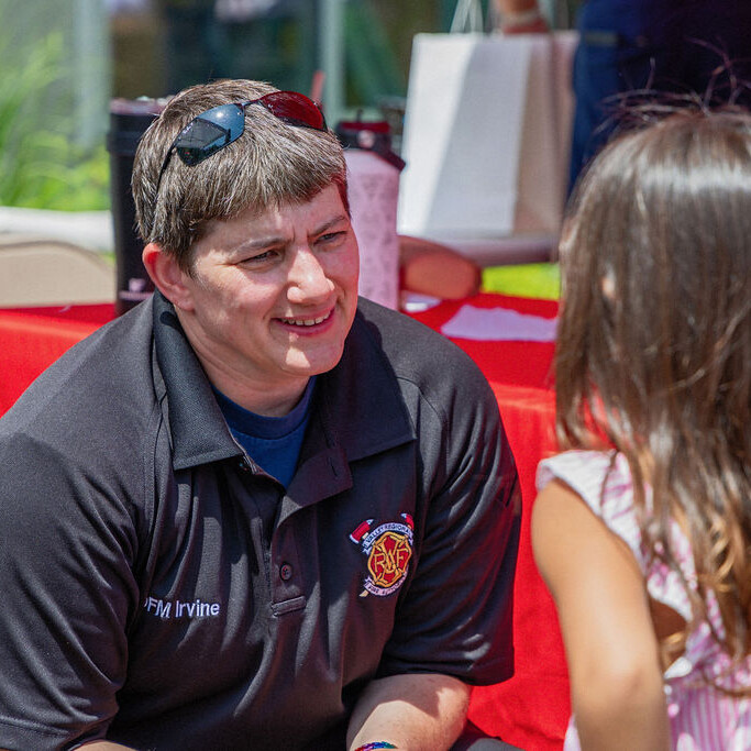 A firefighter in uniform kneels and talks to a young girl at an outdoor community event with tables and red tablecloths.