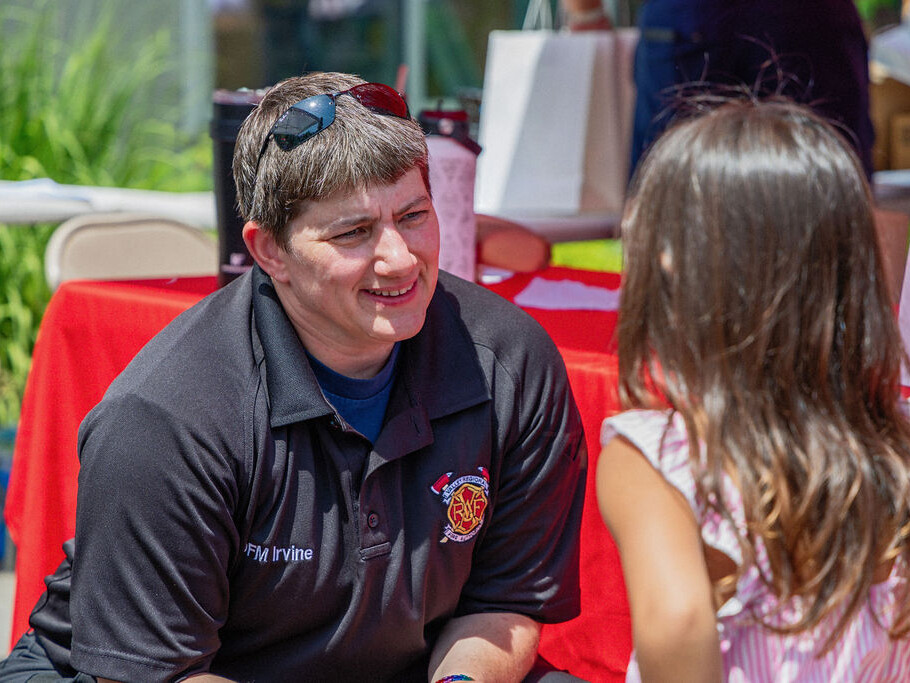 A firefighter in uniform kneels and talks to a young girl at an outdoor community event with tables and red tablecloths.