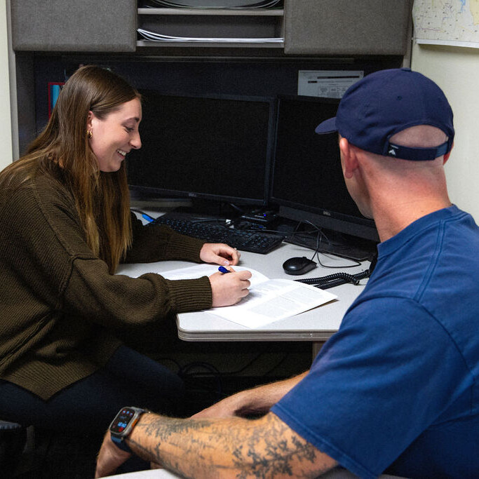 A woman sits at a desk, writing on a document, while a man in a blue shirt and cap sits across from her in an office setting.