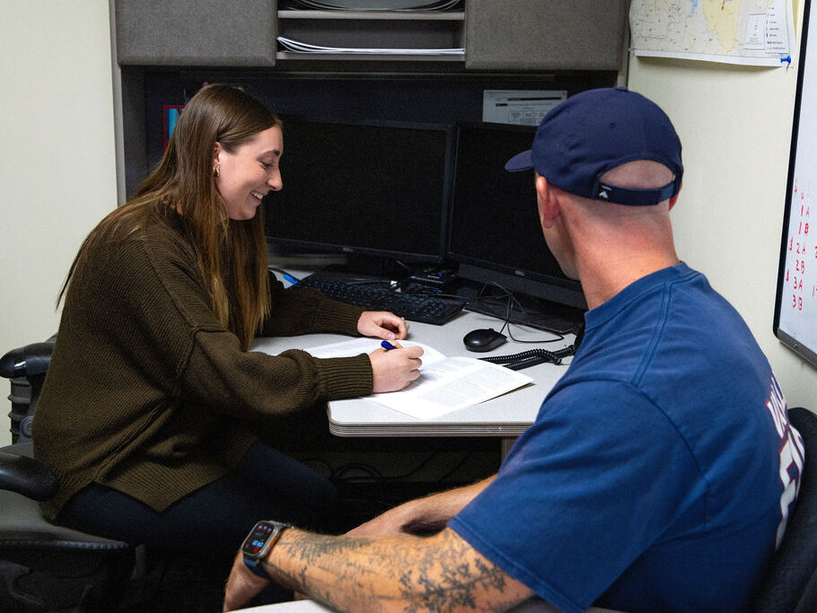 A woman sits at a desk, writing on a document, while a man in a blue shirt and cap sits across from her in an office setting.