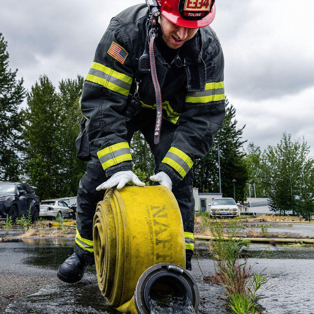 A firefighter in full gear and a red helmet unrolls a yellow fire hose with water flowing out, standing on wet pavement outdoors.