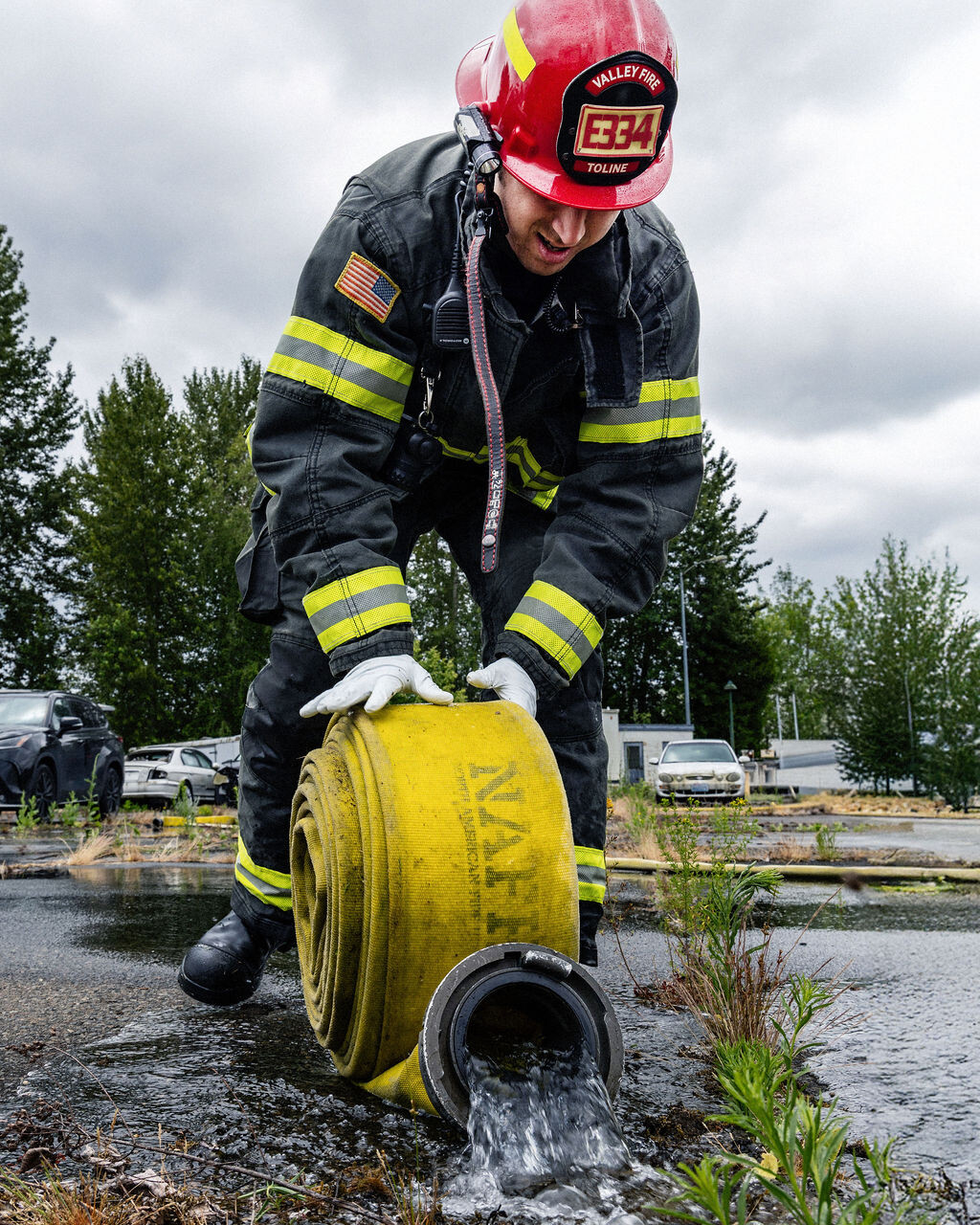 A firefighter in full gear and a red helmet unrolls a yellow fire hose with water flowing out, standing on wet pavement outdoors.