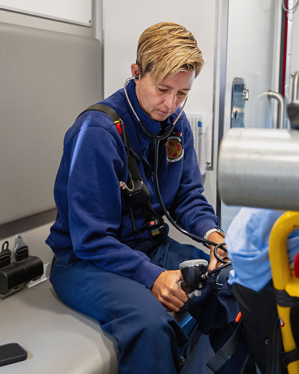 A paramedic wearing a headset and uniform sits on a bench inside an ambulance, preparing or checking medical equipment.