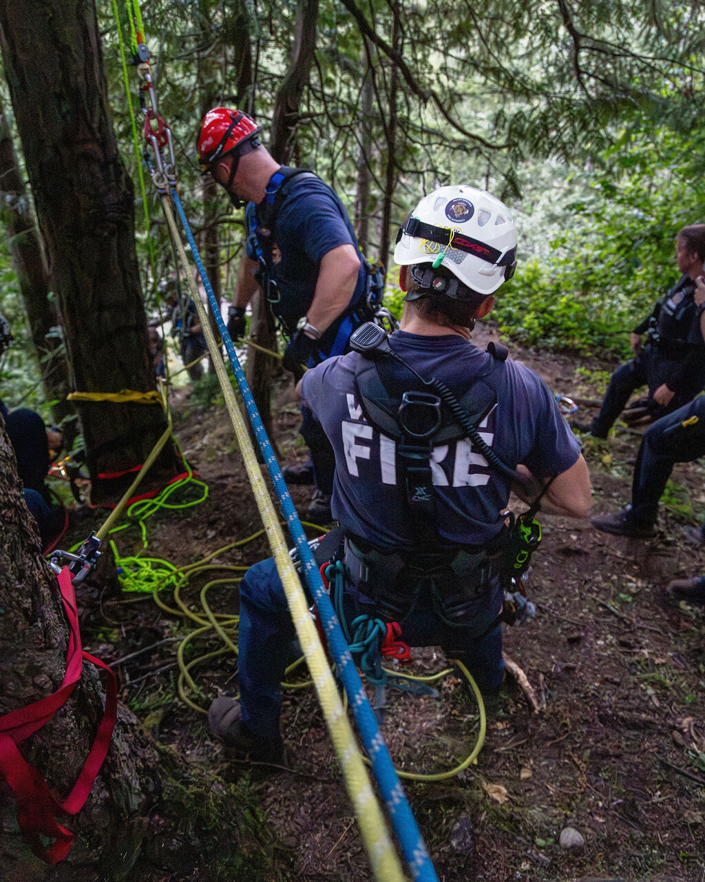 Two rescue workers in helmets and harnesses perform a rope rescue operation in a forested area, with safety lines and gear attached to trees.