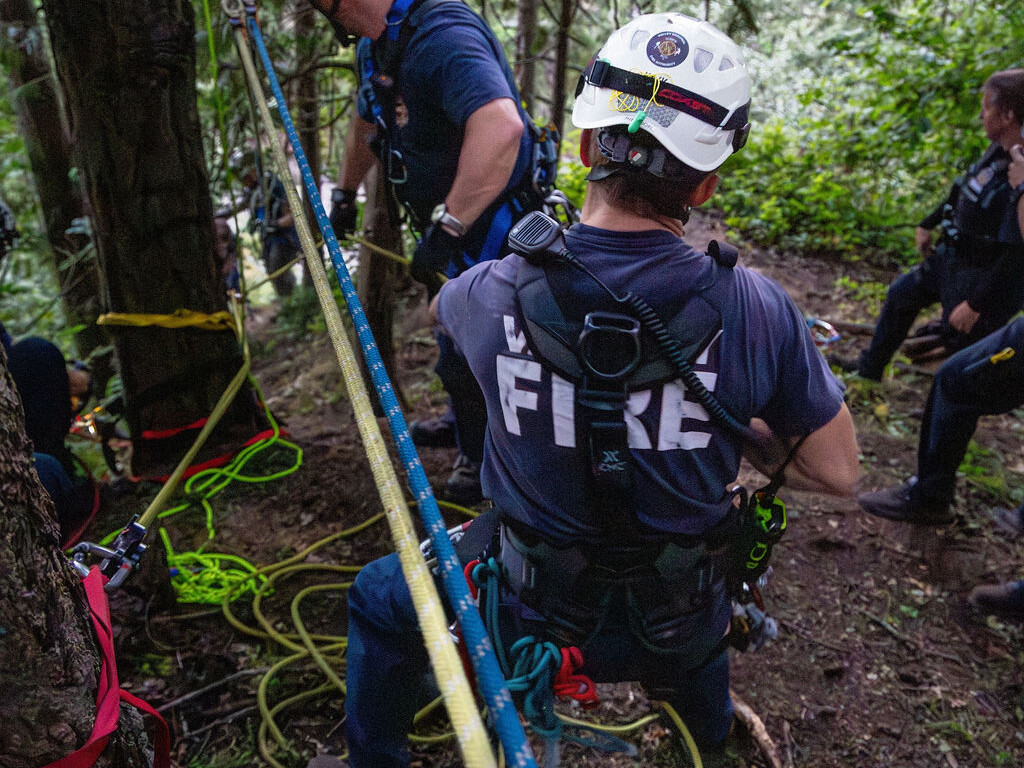 Two rescue workers in helmets and harnesses perform a rope rescue operation in a forested area, with safety lines and gear attached to trees.
