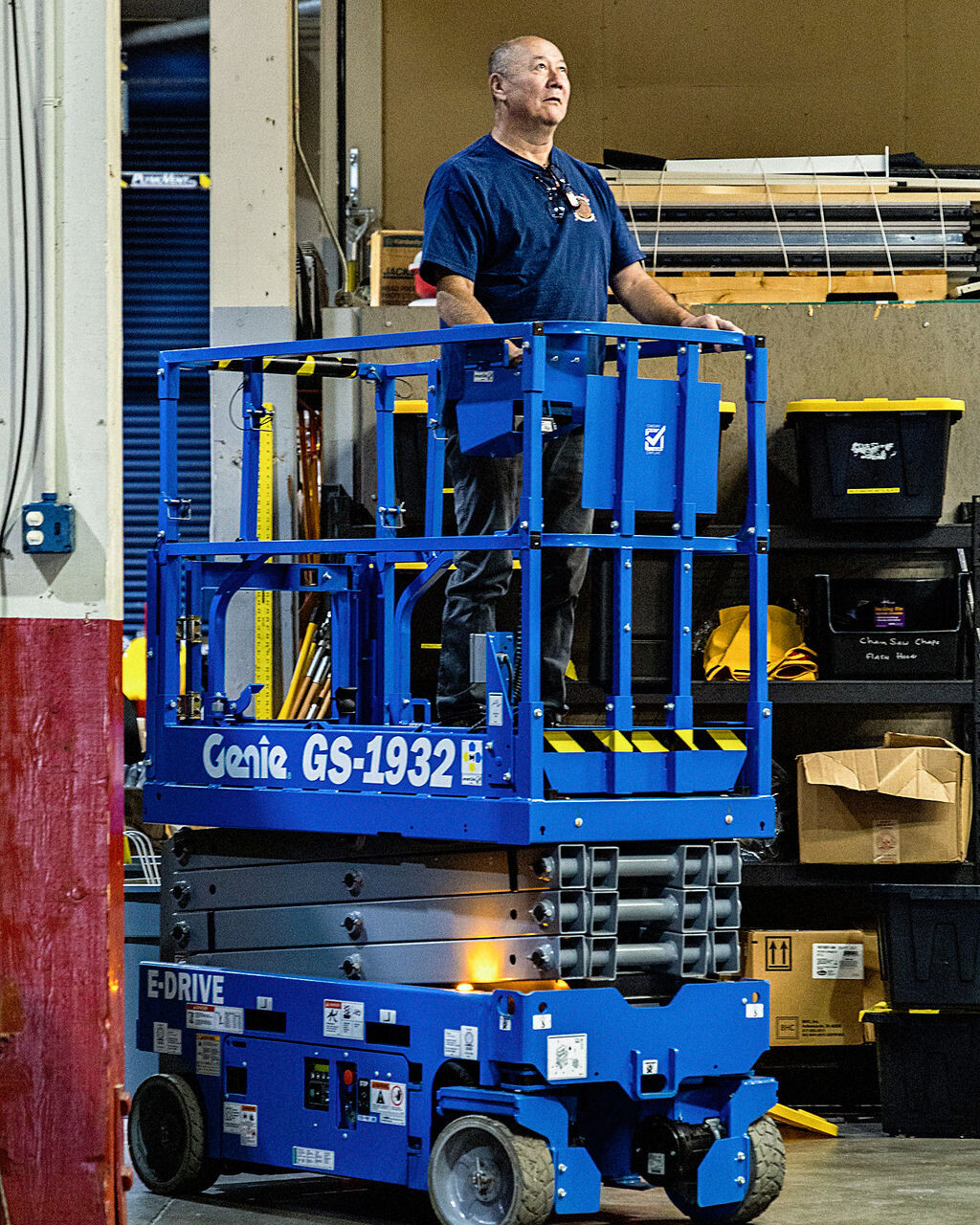 A man stands on a blue Genie GS-1932 scissor lift in an indoor industrial setting with boxes and storage bins in the background.