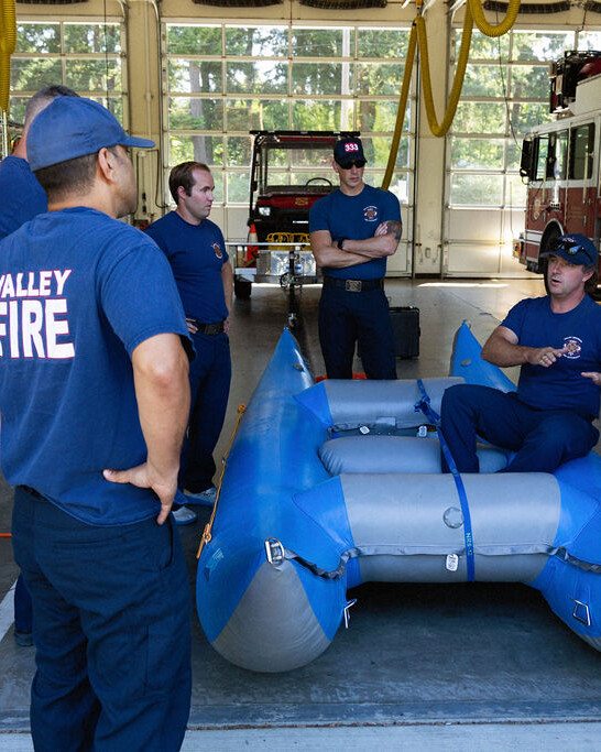 Four firefighters in uniform stand around an inflatable rescue raft in a fire station, while one seated firefighter gives instructions. Fire trucks and equipment are visible in the background.