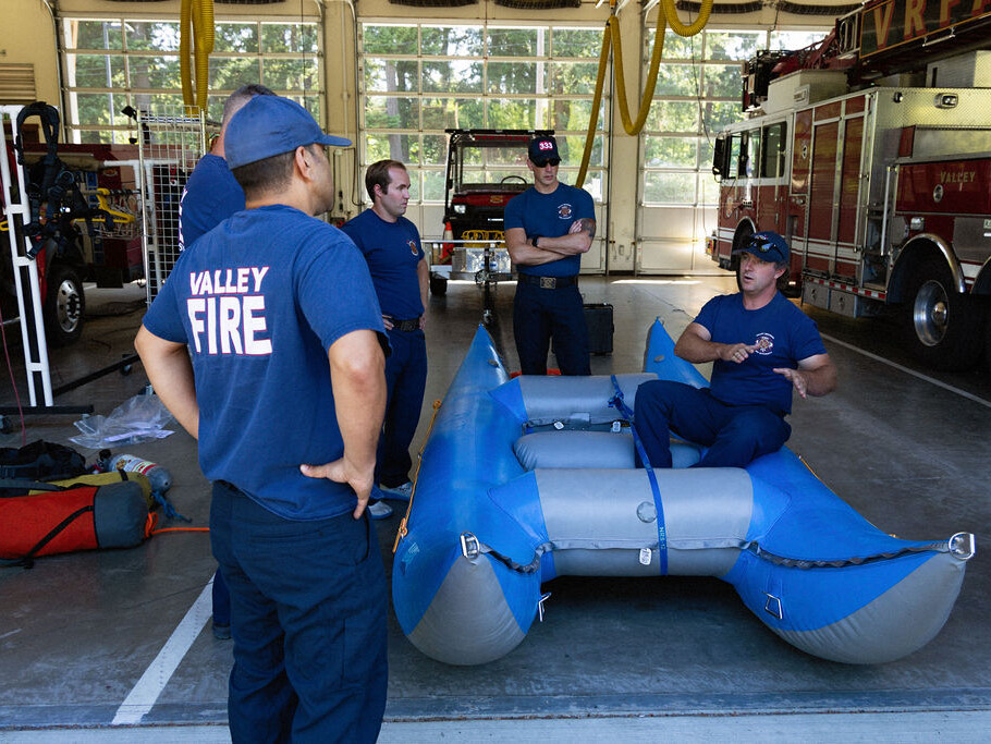 Four firefighters in uniform stand around an inflatable rescue raft in a fire station, while one seated firefighter gives instructions. Fire trucks and equipment are visible in the background.