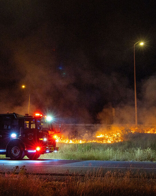 A fire truck and firefighter respond to a grass fire burning beside a road at night, with smoke rising and streetlights illuminating the scene.