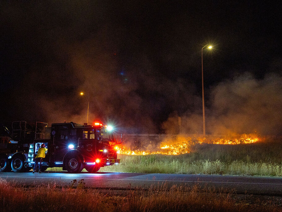 A fire truck and firefighter respond to a grass fire burning beside a road at night, with smoke rising and streetlights illuminating the scene.