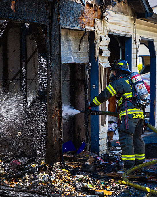 A firefighter sprays water on the charred remains of a building while a fire truck is parked nearby. Burnt debris is scattered on the ground.