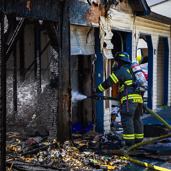 A firefighter sprays water on the charred remains of a building while a fire truck is parked nearby. Burnt debris is scattered on the ground.