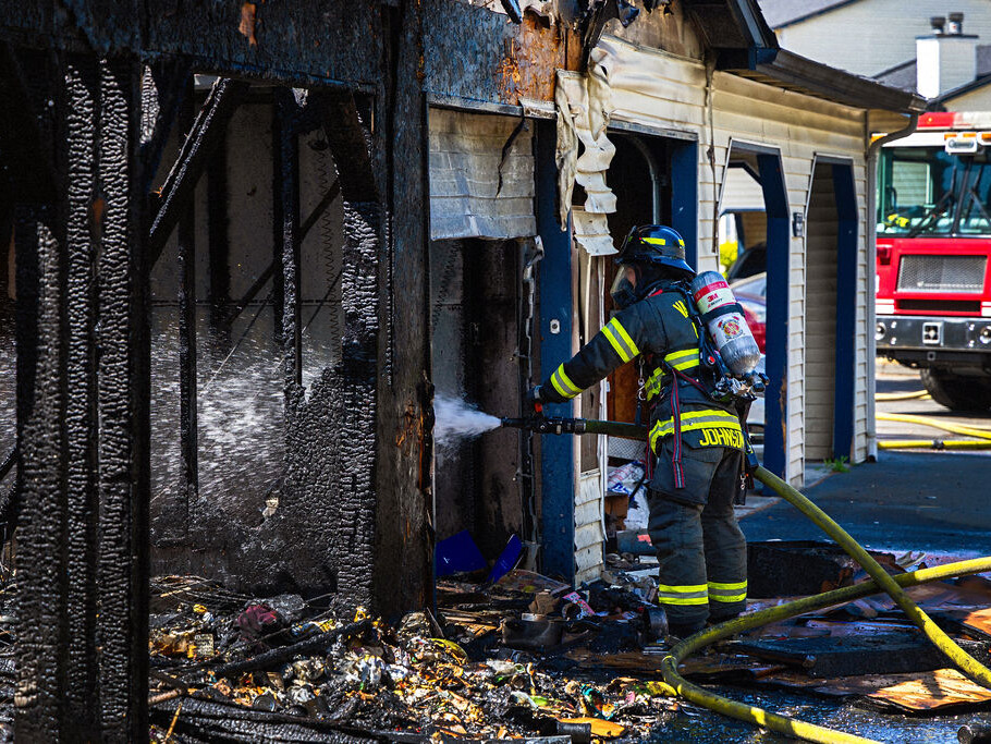 A firefighter sprays water on the charred remains of a building while a fire truck is parked nearby. Burnt debris is scattered on the ground.