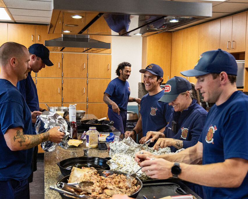 Six firefighters in uniform prepare food together in a kitchen, wrapping items in foil and working around the counter with various ingredients.