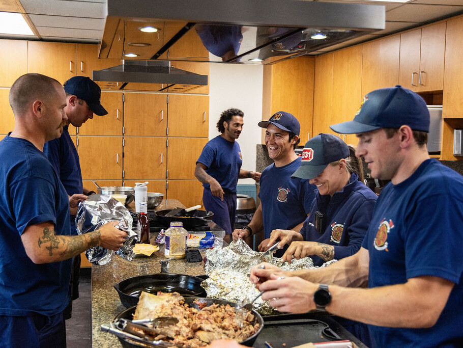 Six firefighters in uniform prepare food together in a kitchen, wrapping items in foil and working around the counter with various ingredients.