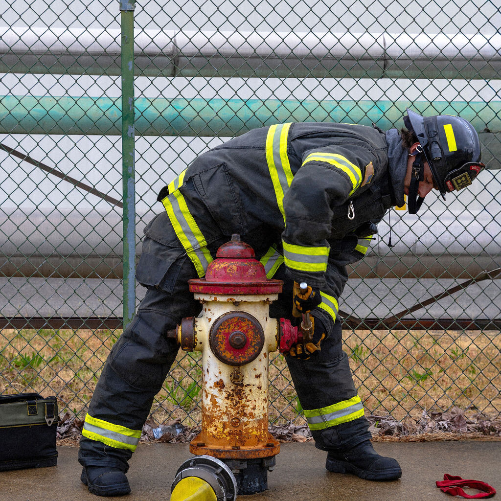 A firefighter in full gear connects a yellow hose to a rusty fire hydrant on a wet sidewalk near a chain-link fence.