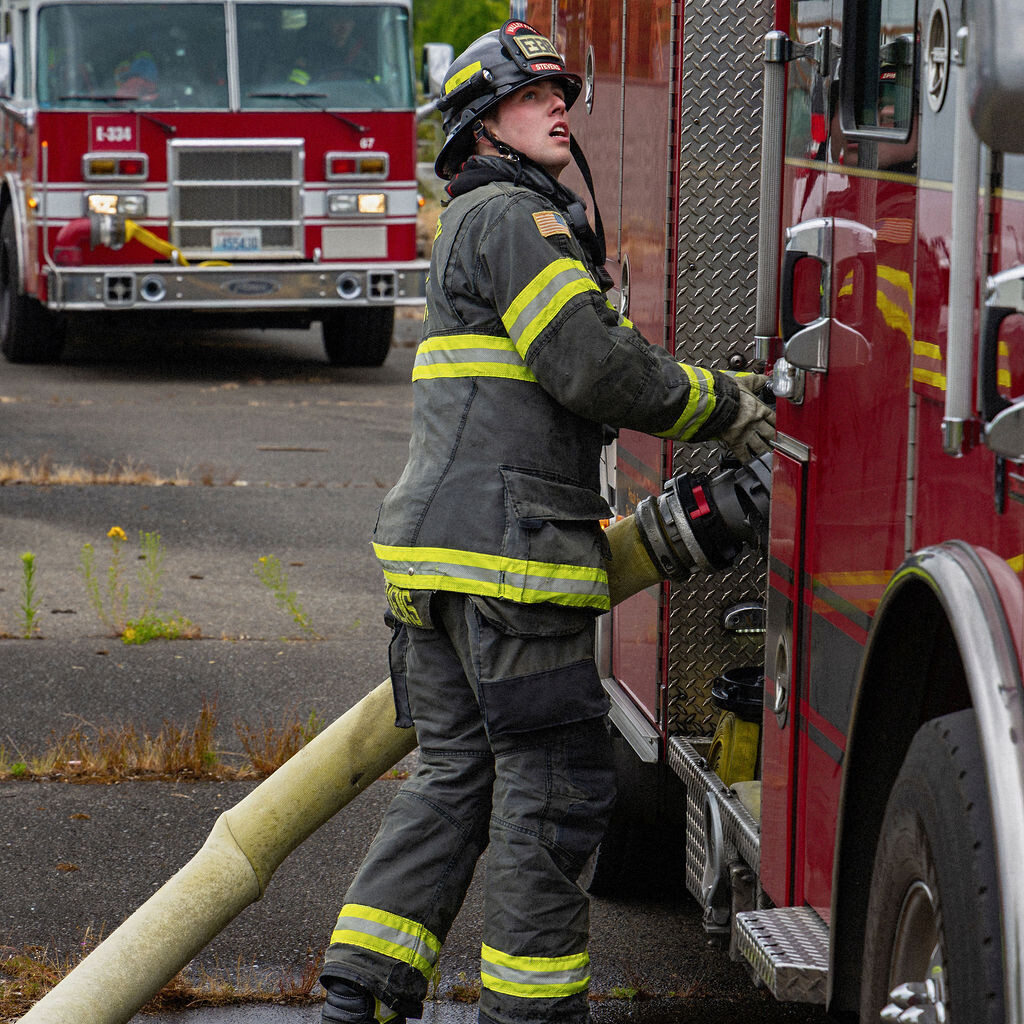 A firefighter in full gear connects a large hose to a fire truck, with another fire truck visible in the background on a wet pavement.