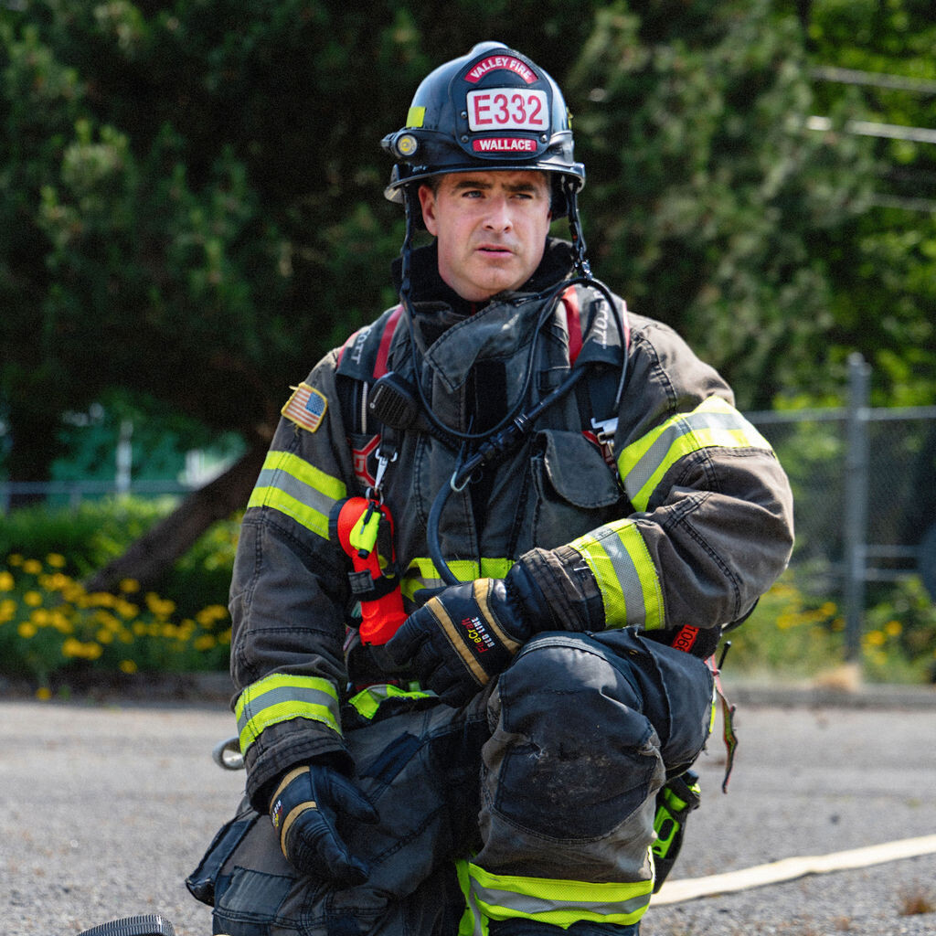 A firefighter in full gear kneels on the ground outdoors, holding a fire hose with a nozzle. Trees and a fence are visible in the background.