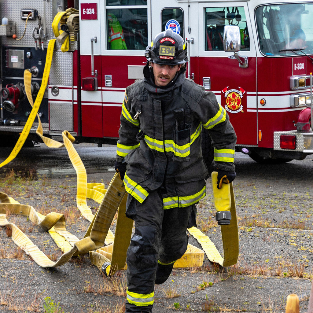 A firefighter in full gear drags a fire hose near a red fire truck parked on a gravel lot.