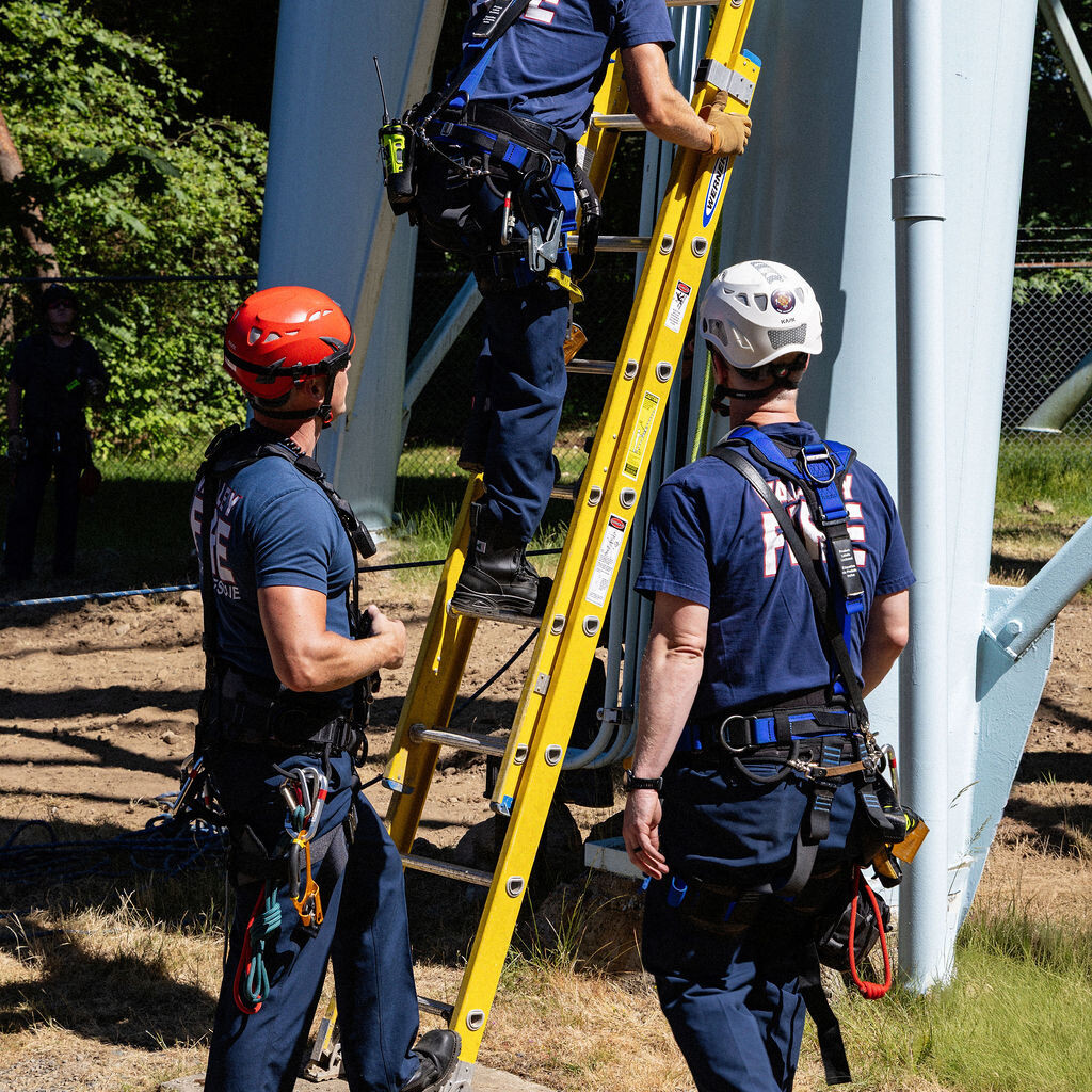 Three rescue workers in helmets and harnesses stand at the base of a yellow ladder, with one worker climbing up towards a metal structure outdoors.
