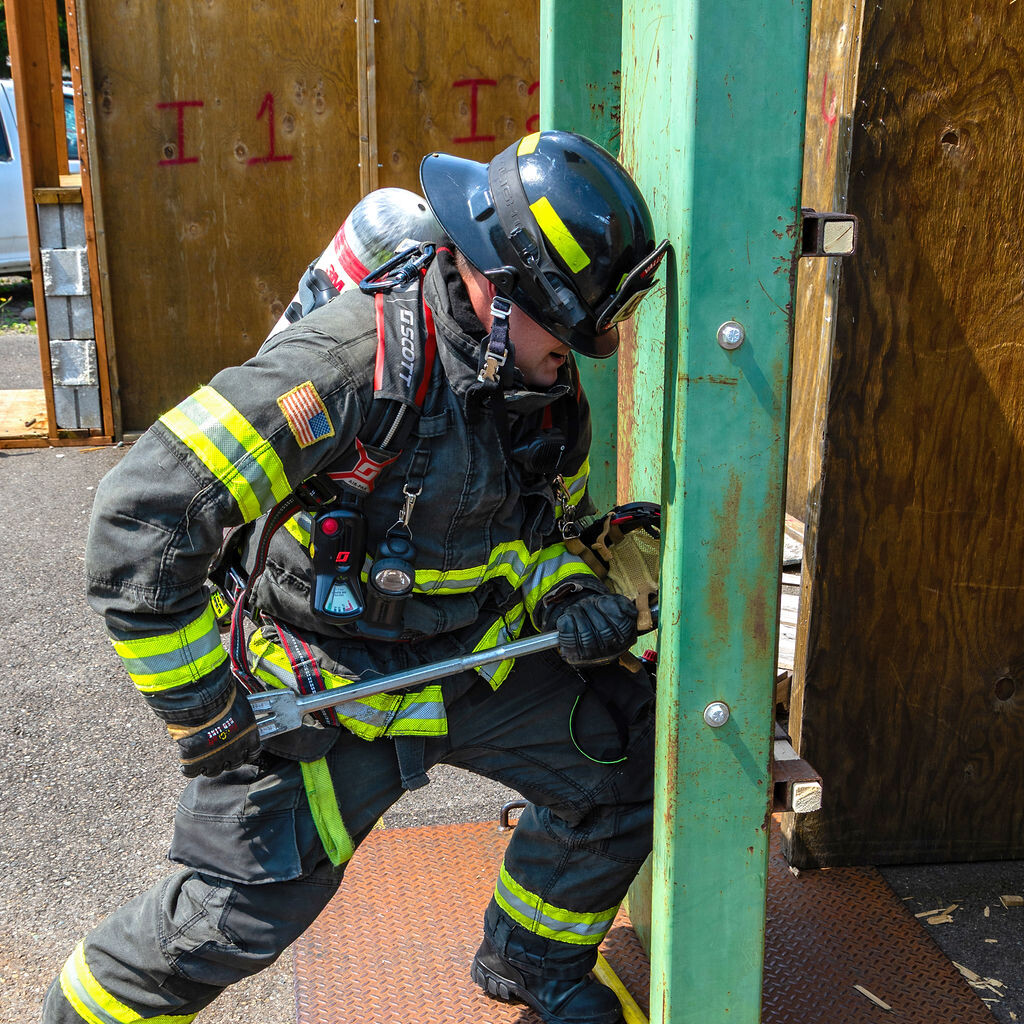 A firefighter in full gear uses a tool to apply force to a green metal structure during a training exercise.