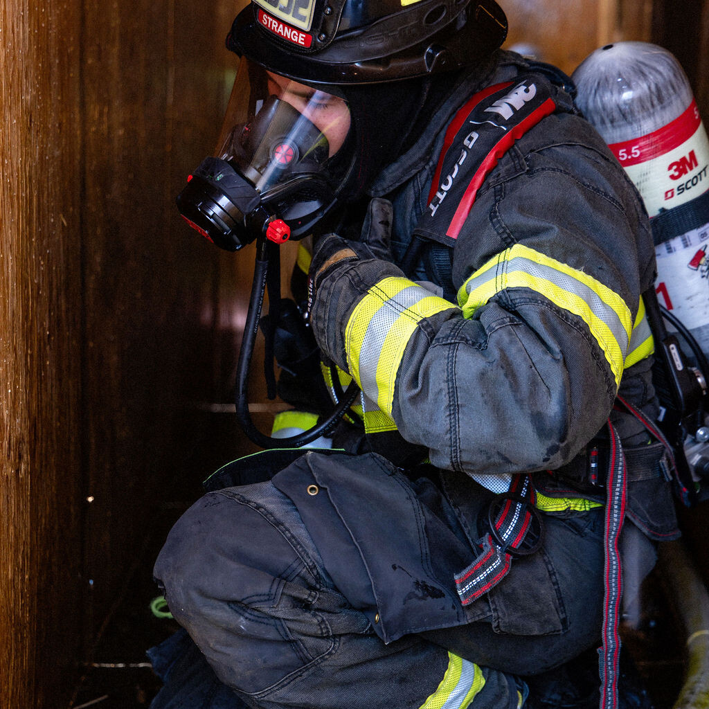 A firefighter in full gear and breathing apparatus crouches in a confined, wooden space holding a hose.