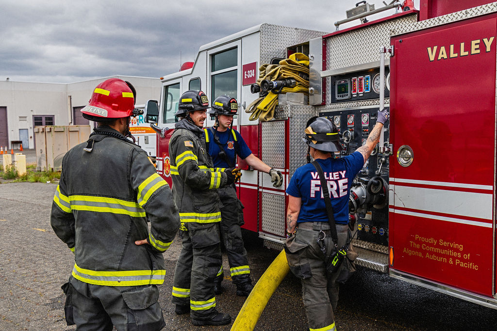 Four firefighters in gear stand next to a fire truck, with one adjusting controls on the side panel. Coiled hoses and equipment are visible on the truck.