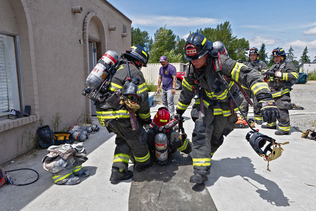 Firefighters in full gear practice a rescue drill, carrying a person on the ground near a building while others observe and standby.