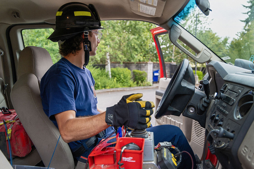 A firefighter wearing gloves and a helmet sits in the driver's seat of a vehicle, operating equipment with one hand while looking outside through the open door.