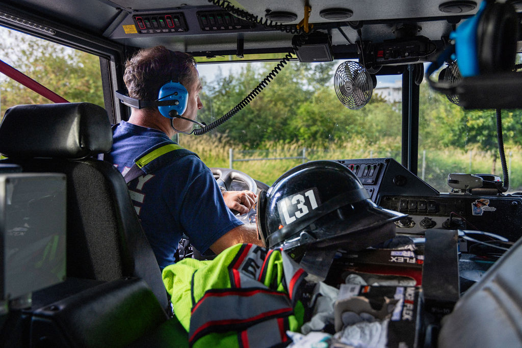 A firefighter wearing a headset sits in the driver’s seat of a fire truck. A helmet labeled “L37” and safety gear are on the dashboard. Trees are visible through the windshield.