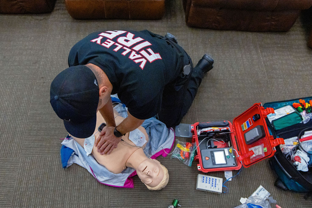 A firefighter performs CPR on a training mannequin next to medical equipment on the floor.