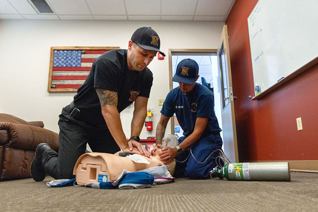 Two firefighters practice CPR and use a resuscitator mask on a training mannequin in a room with an American flag on the wall.