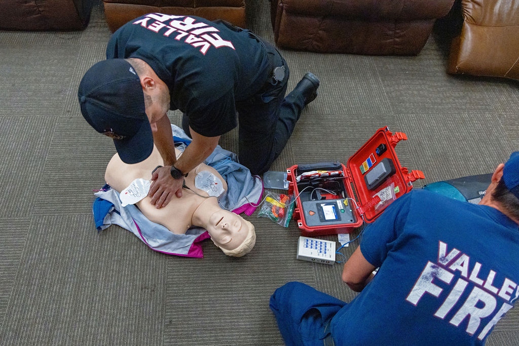 Two firefighters in uniforms practice CPR on a training manikin with defibrillator pads, next to medical equipment on the floor.