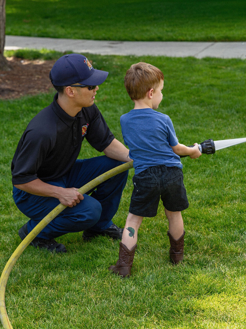 A firefighter kneels on grass, helping a young boy use a fire hose. The boy wears a blue shirt, black shorts, and cowboy boots.